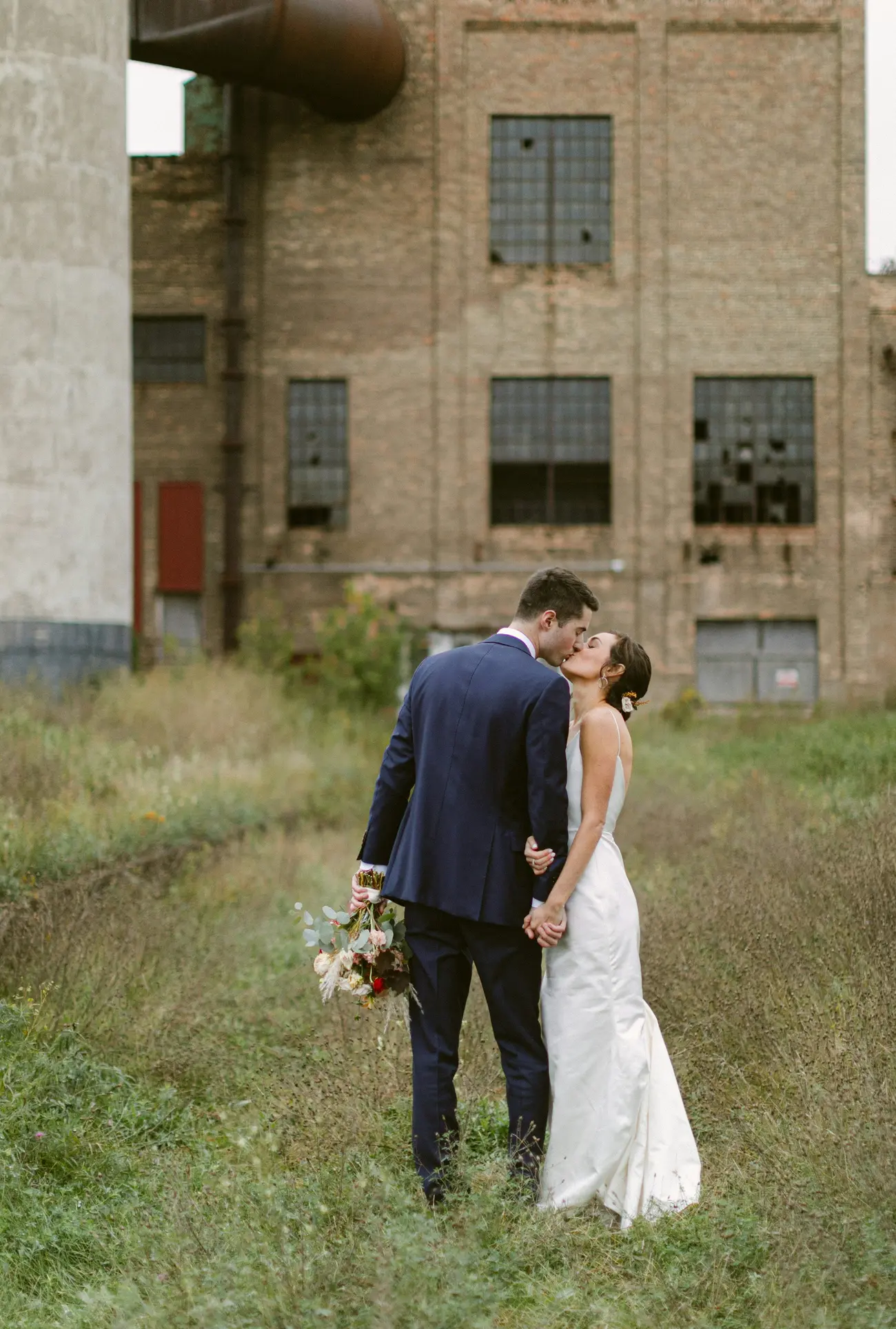 A couple kissing in tall grass beside the depot's brick wall and tall factory windows