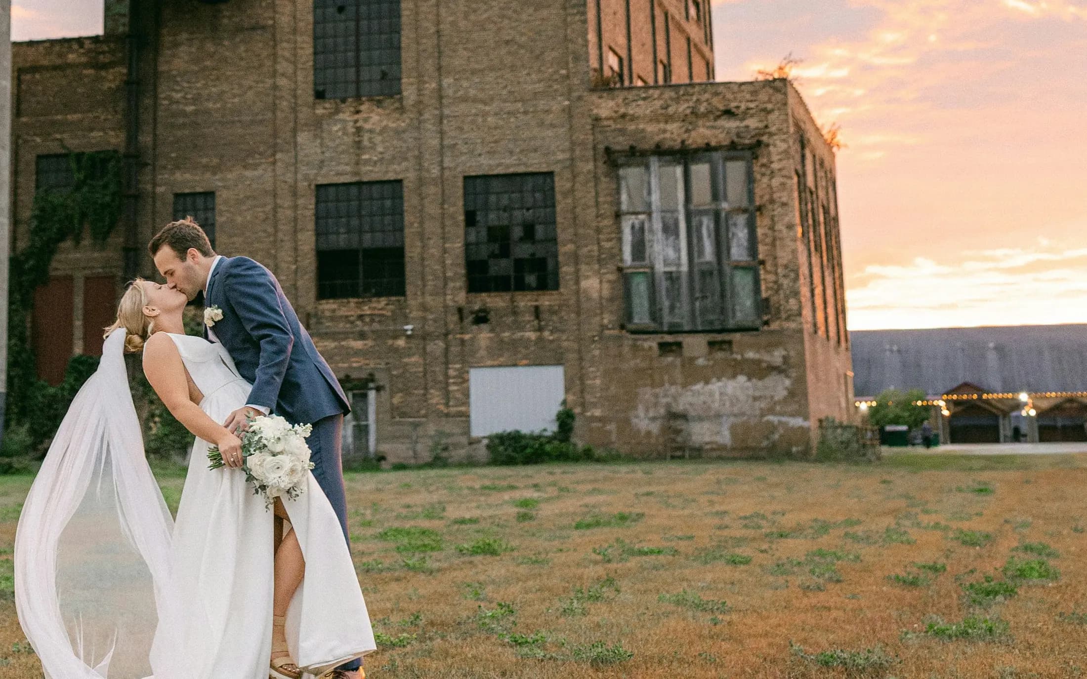 A bride and groom kissing on the grass in front of the old brick depot at sunset, with the lit Platform roof to the right