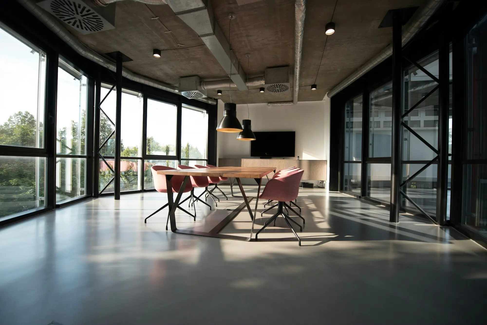 A finished corner office floor with steel-framed windows on two sides, a table set under pendant lights and spotless polished concrete
