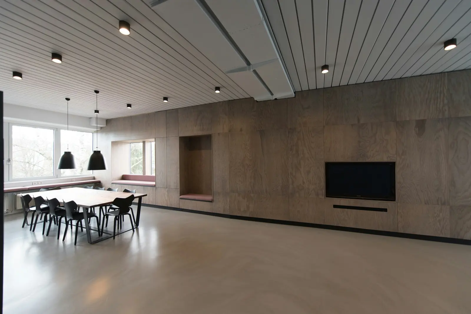 An empty office lounge at first light: plywood wall, a long table with chairs pushed in, a spotless pale floor