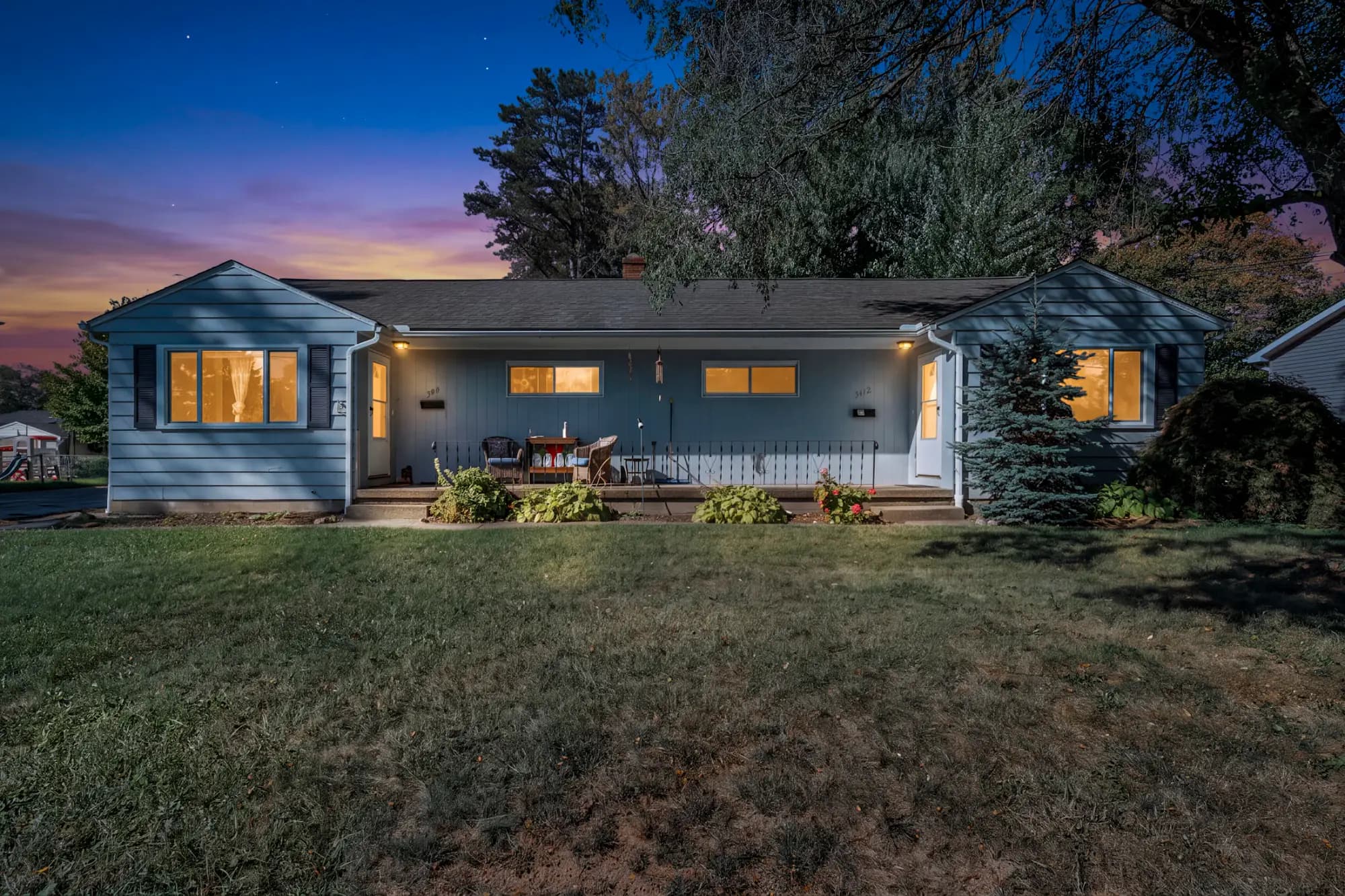 A single-story ranch house at dusk, porch lights and windows lit, on a wide front lawn
