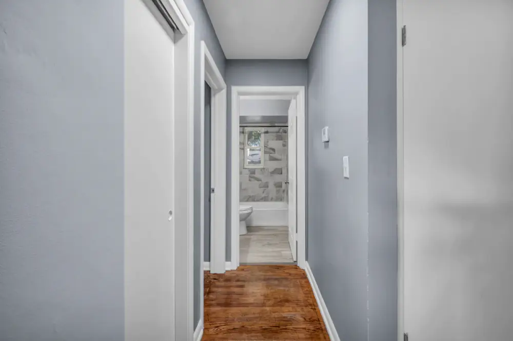 A narrow hallway with light blue walls and oak floors leading to a tiled bathroom