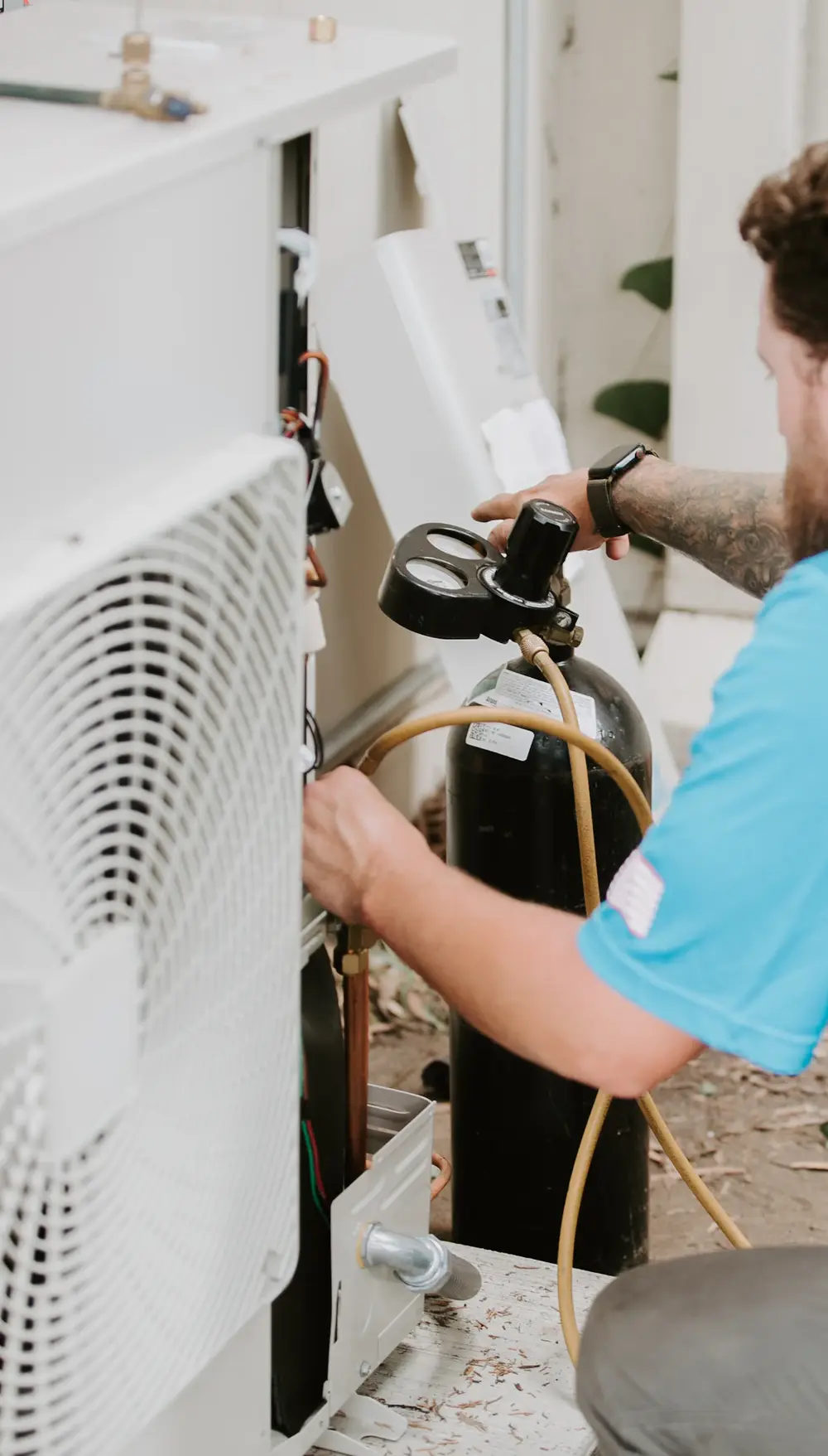 A technician kneeling at an outdoor AC unit with refrigerant gauges and a nitrogen tank