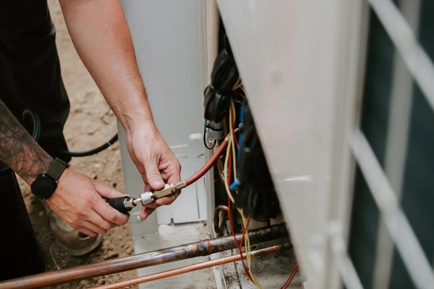 A technician's hands connecting a refrigerant hose to the service valve of an outdoor unit, copper lines below
